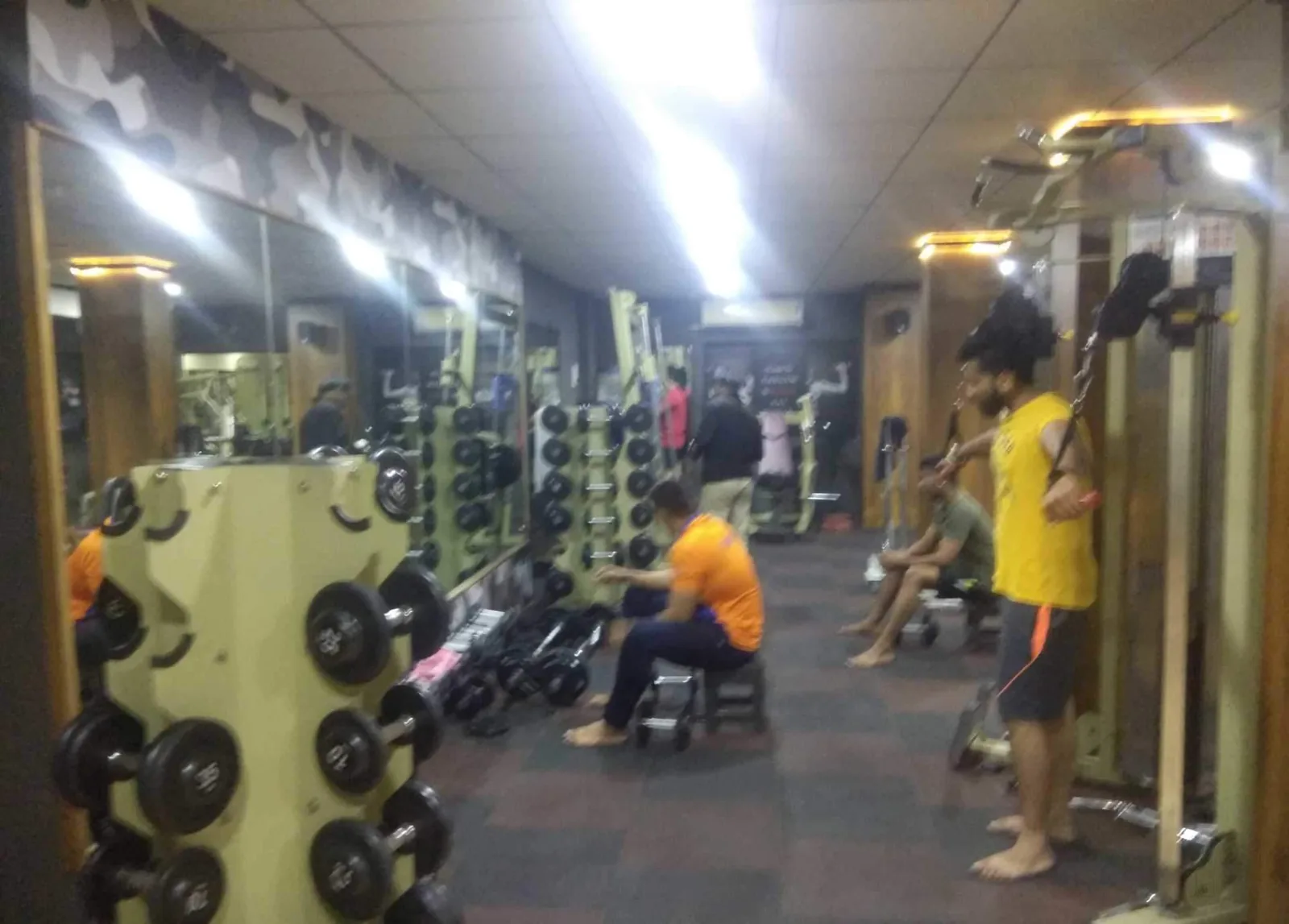Members training on the floor at S R Gym Katargam, Surat, with dumbbell racks along both walls and a cable machine in use