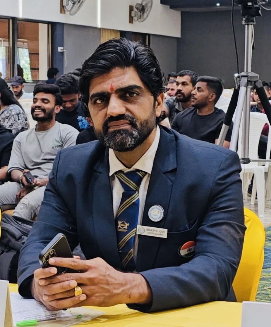 Brijesh Joshi seated at an officials' table at a bodybuilding championship, wearing a badge reading BRIJESH S. JOSHI SOUTH GUJARAT
