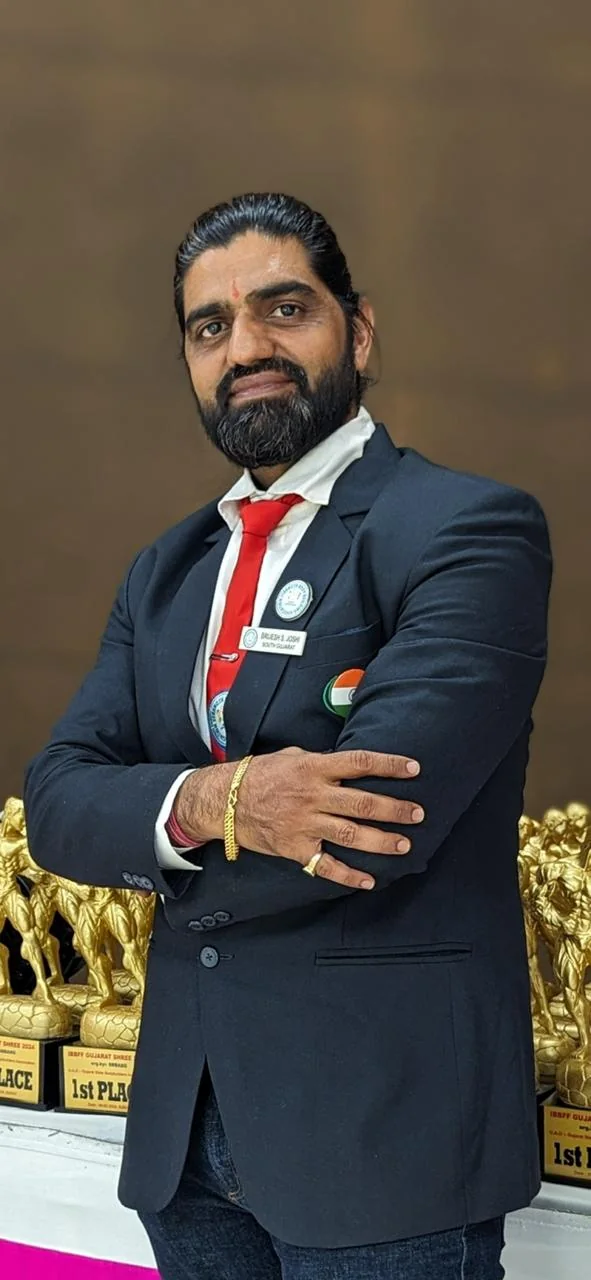 Brijesh Joshi in an official association blazer with a South Gujarat name badge, standing in front of IBBFF Gujarat Shree first-place trophies