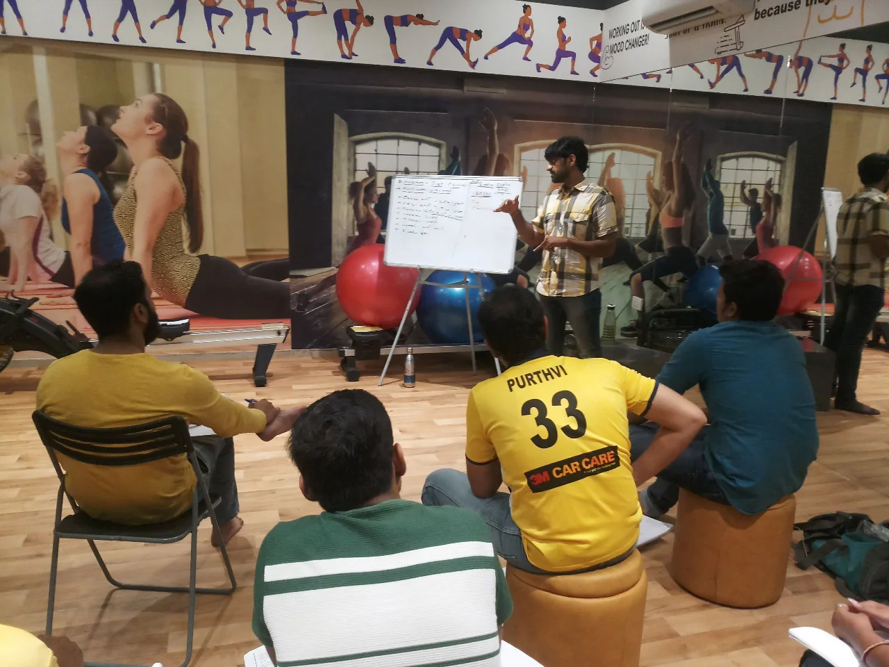 Brijesh Joshi teaching at a whiteboard to a seated group of trainee fitness instructors on a gym floor in Surat