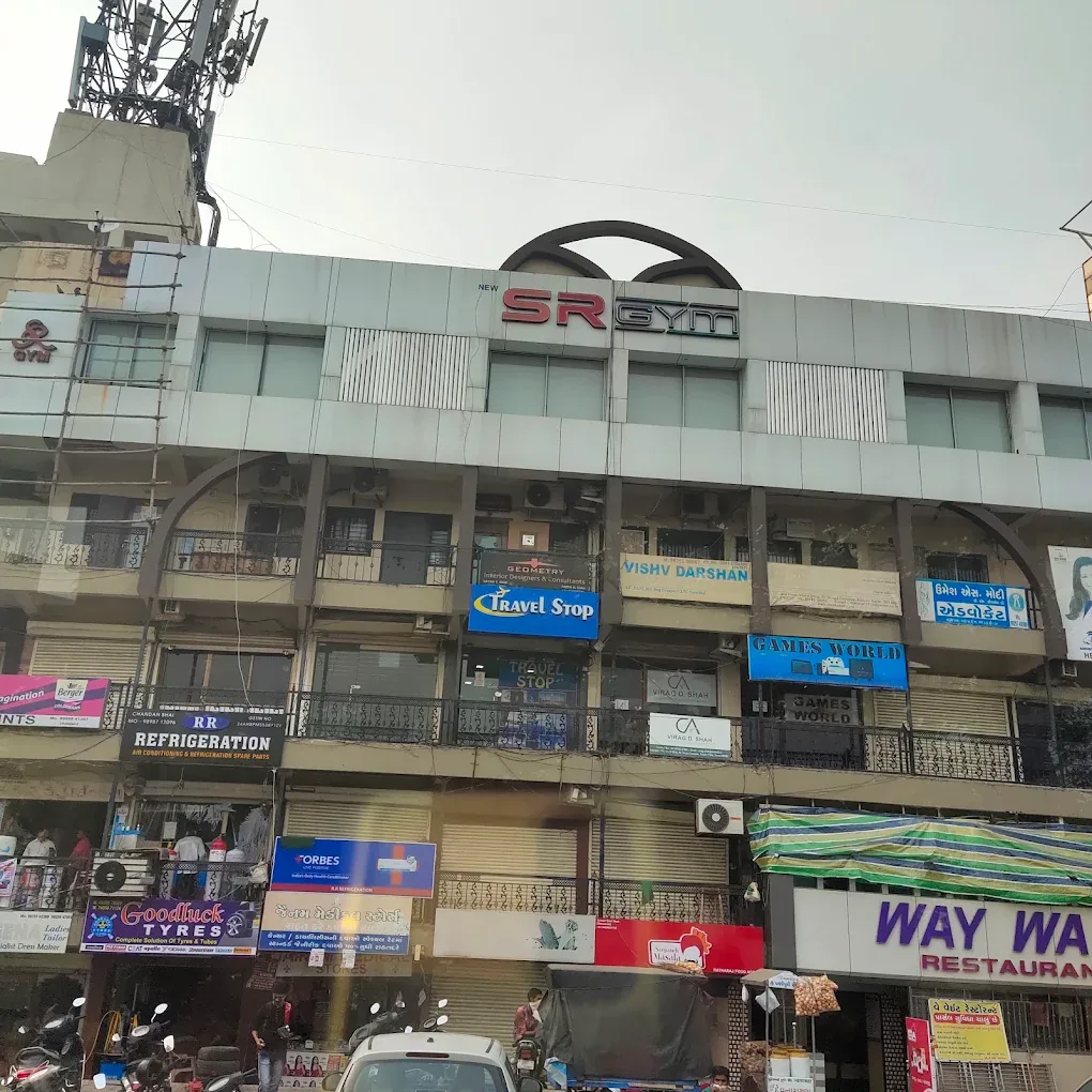 The street frontage of the building housing S R Gym Adajan in Surat. The red and grey 'SR GYM' sign sits under a curved arch on the top floor, above Travel Stop and Games World on the middle floor and Way Wait Restaurant at street level.