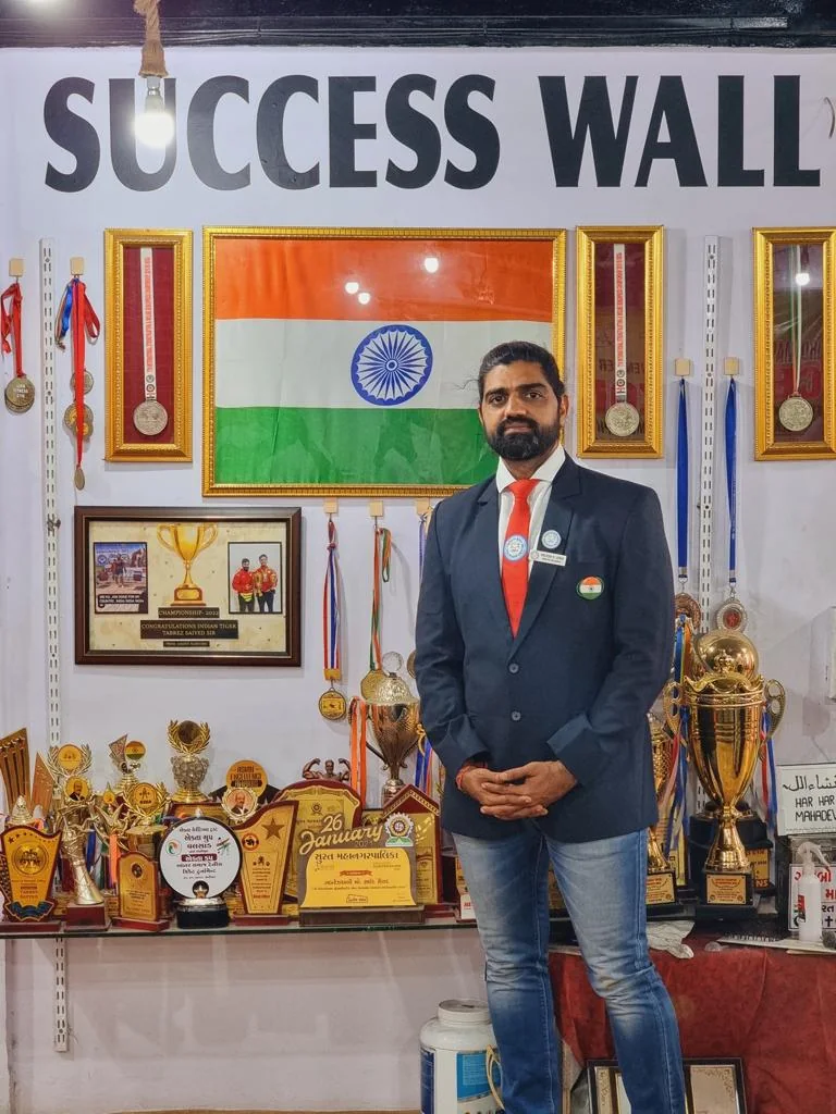 Brijesh Joshi in his association blazer in front of the success wall at S R Gym, hung with members' medals, trophies and championship certificates