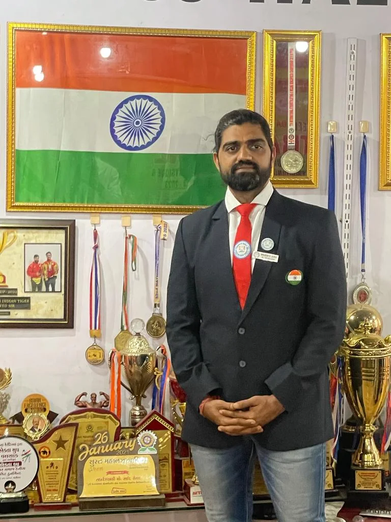 Brijesh Joshi in an association blazer standing in front of a wall of bodybuilding trophies, medals and framed certificates at S R Gym, Surat