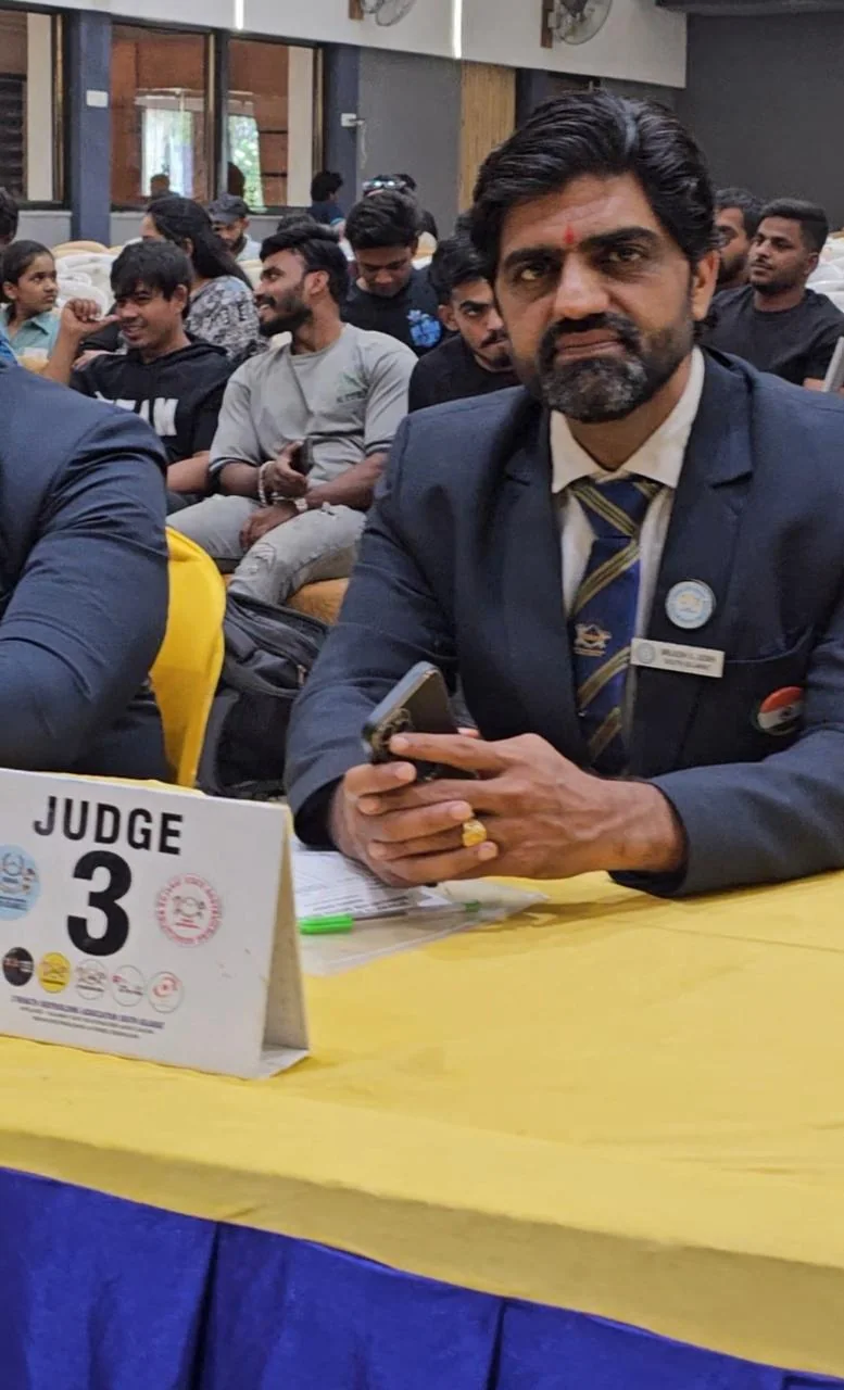 Brijesh Joshi seated at a judging table marked Judge 3 at a bodybuilding championship